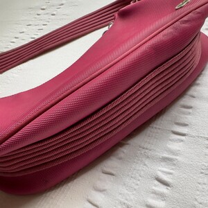 May include: A bright pink handbag with a textured surface and a ribbed shoulder strap. The bag has a curved shape and a pleated design on the bottom. The strap is also pink and ribbed.