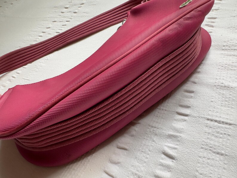 May include: A bright pink handbag with a textured surface and a ribbed shoulder strap. The bag has a curved shape and a pleated design on the bottom. The strap is also pink and ribbed.