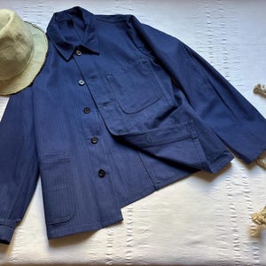 Könnte beinhalten: Marineblaue Arbeitsjacke mit Knopfleiste und zwei Vordertaschen. Ein Strohhut und ein Seil sind ebenfalls im Bild. Die Jacke hat ein Fischgrätenmuster.