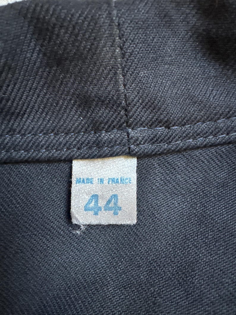 May include: Close-up of a dark gray fabric with a stitched seam. A small, rectangular tag is sewn onto the fabric, displaying the text "MADE IN FRANCE" above the number "44" in blue.