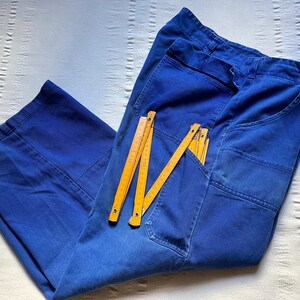 May include: A pair of blue work pants featuring reinforced knee panels and multiple pockets. A yellow folding wooden ruler is placed on top of the pants, showing measurements in centimeters up to 200 cm, with the 50 cm mark clearly visible.