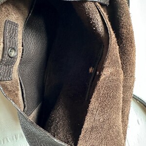 May include: A dark brown leather shoulder bag with a textured exterior. The interior is a lighter brown suede. The bag has a snap closure and a shoulder strap. The bag is open, revealing the interior.