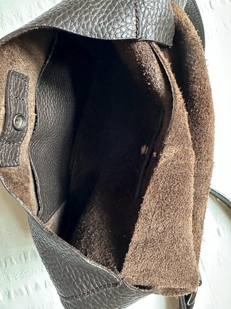 May include: A dark brown leather shoulder bag with a textured exterior. The interior is a lighter brown suede. The bag has a snap closure and a shoulder strap. The bag is open, revealing the interior.