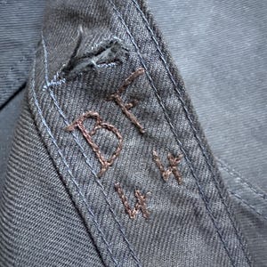 May include: Close-up of a section of dark blue denim fabric. The fabric features visible stitching in a lighter blue thread along the seams. Embroidered in brown thread are the letters "BF" and the numbers "14 14".