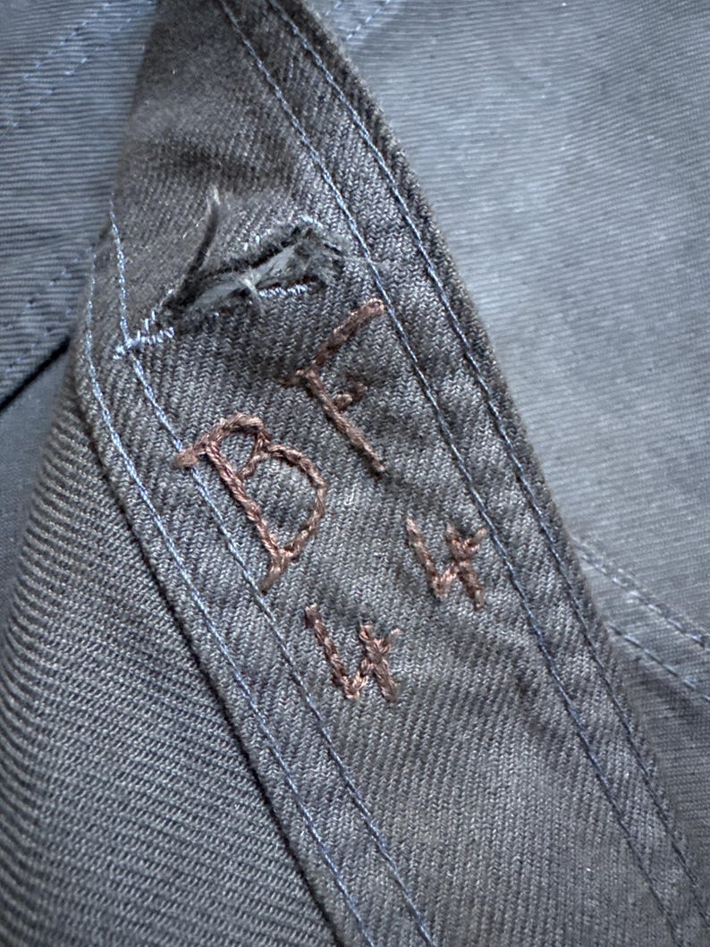 May include: Close-up of a section of dark blue denim fabric. The fabric features visible stitching in a lighter blue thread along the seams. Embroidered in brown thread are the letters "BF" and the numbers "14 14".