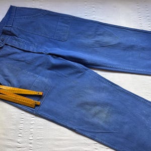 May include: A pair of blue denim work pants with a large pocket on the right leg and a smaller pocket on the left leg. The pants have a button fly and a belt loop. There are four wooden folding rulers in the pocket.