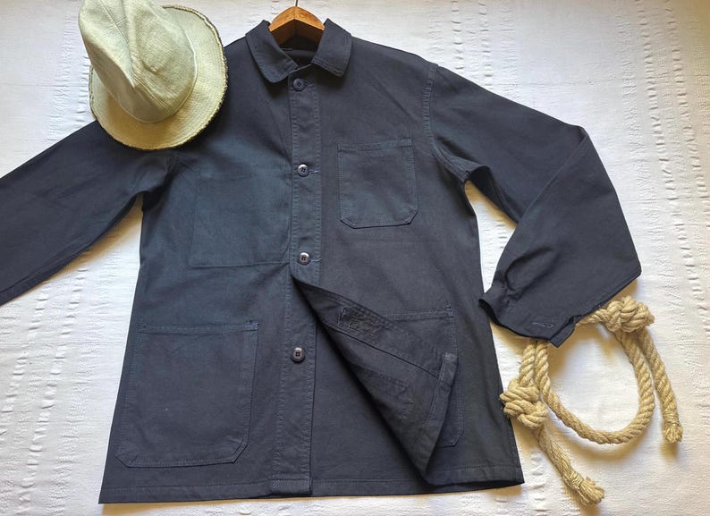 May include: A dark navy blue work jacket with a button-up front, collar, and multiple pockets. A light green straw hat rests on the jacket's shoulder. A length of rope with knots is draped near the jacket.