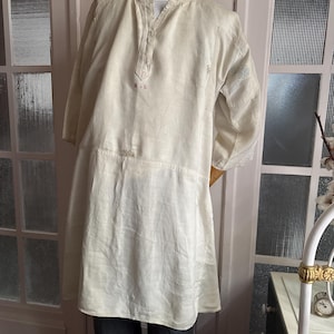Op de afbeelding: Crèmekleurige tuniek met een opstaande kraag en lange mouwen. De tuniek heeft een relaxte pasvorm en een knoopsluiting. De stof lijkt linnen of een vergelijkbaar lichtgewicht materiaal te zijn. Er is een kleine rode markering op de borst.