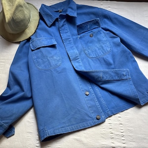 Op de afbeelding: Een blauwe denim werkjas met twee grote zakken en een knoopsluiting. De jas heeft een versleten look en is gemaakt van een stevige stof.
