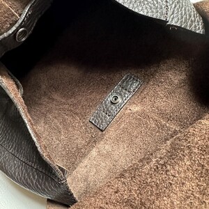 May include: A dark brown leather tote bag with a textured exterior and a soft, suede-like interior. The bag features a snap closure and a spacious open design, perfect for everyday use.