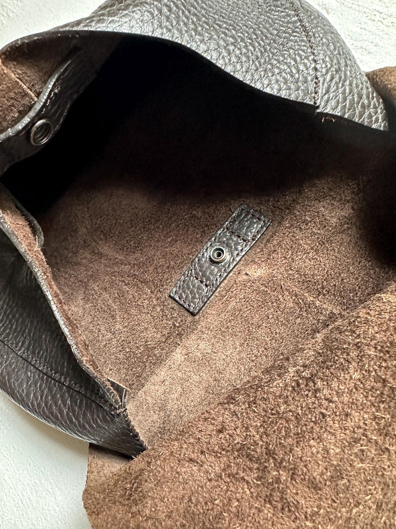 May include: A dark brown leather tote bag with a textured exterior and a soft, suede-like interior. The bag features a snap closure and a spacious open design, perfect for everyday use.