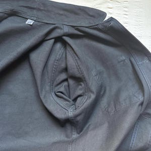 May include: Close-up of a dark gray jacket with a visible pocket and stitching details. The jacket is made of a sturdy fabric, and the size tag reads "42". The overall style suggests a workwear or utility garment.