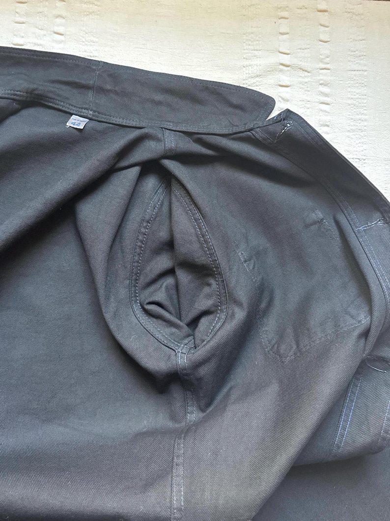 May include: Close-up of a dark gray jacket with a visible pocket and stitching details. The jacket is made of a sturdy fabric, and the size tag reads "42". The overall style suggests a workwear or utility garment.