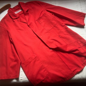 May include: Bright red lab coat with three front pockets and a notched collar. The coat has long sleeves and a button closure. A wooden hanger is visible in the upper left corner.