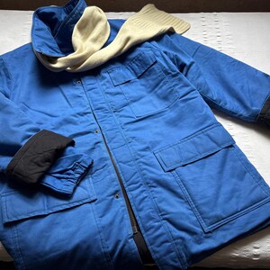 May include: A blue winter jacket with a black lining and a white scarf. The jacket has a zipper closure and multiple pockets.