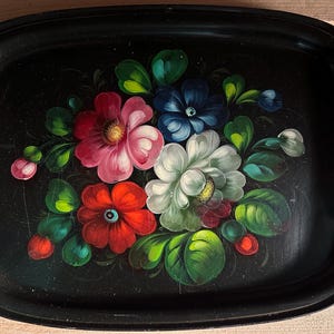 Può includere: Vassoio rettangolare nero con un motivo floreale dipinto a mano. I fiori sono nelle tonalità del rosso, rosa, bianco e blu, con foglie verdi. Il vassoio ha una finitura lucida e un aspetto leggermente usurato.