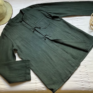 May include: A dark green long-sleeved shirt with a mandarin collar and a single pocket on the left chest. The shirt has a tie closure at the neck.