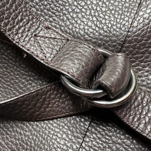 May include: Close-up of a dark brown leather bag with a textured surface. A silver metal ring connects the strap to the bag. The leather has a pebbled grain.