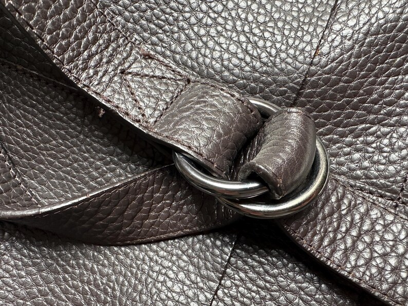 May include: Close-up of a dark brown leather bag with a textured surface. A silver metal ring connects the strap to the bag. The leather has a pebbled grain.