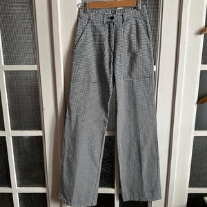 May include: High-waisted trousers featuring a black and white micro-check pattern. These pants include a front button closure, a zipper fly, two side pockets, and two patch pockets on the front. The leg cut is straight, providing a structured fit.