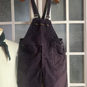 May include: A pair of dark purple overalls with adjustable straps and multiple pockets. The overalls are hanging on a wooden hanger.