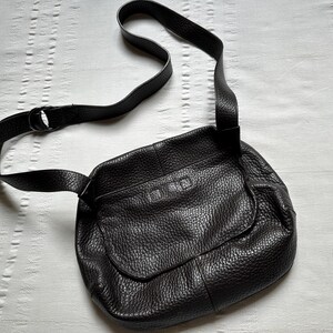 May include: A black leather crossbody bag with a textured surface. The bag has a flap closure and an adjustable shoulder strap. The strap is attached to the bag with a metal ring. The bag is lying on a white surface.