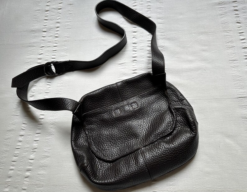 May include: A black leather crossbody bag with a textured surface. The bag has a flap closure and an adjustable shoulder strap. The strap is attached to the bag with a metal ring. The bag is lying on a white surface.