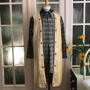 May include: A long, cream-colored vest with a button closure. The vest is worn over a plaid shirt with a bandana tied around the collar. The vest has a long, flowing shape and is made of a soft, fuzzy material.