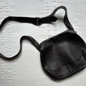 May include: A black leather crossbody bag with a textured surface. The bag has a flap closure and an adjustable strap. The strap is connected to the bag with a metal ring. The bag is lying on a white surface.