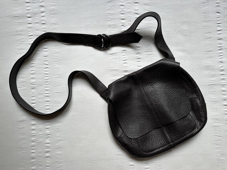 May include: A black leather crossbody bag with a textured surface. The bag has a flap closure and an adjustable strap. The strap is connected to the bag with a metal ring. The bag is lying on a white surface.
