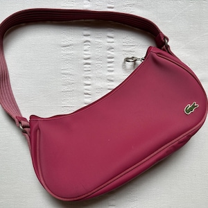 May include: A magenta shoulder bag with a curved design and a matching strap. The bag features a small green crocodile logo on the front. The strap is a lighter shade of magenta. The bag is made of a textured material.
