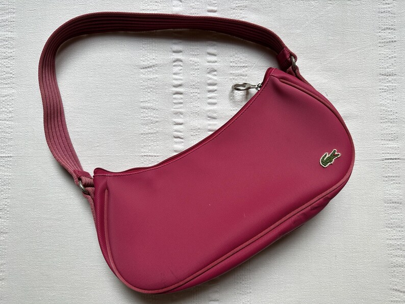 May include: A magenta shoulder bag with a curved design and a matching strap. The bag features a small green crocodile logo on the front. The strap is a lighter shade of magenta. The bag is made of a textured material.