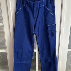 Peut inclure: Pantalon de travail bleu roi avec plusieurs poches et des renforts aux genoux. Le pantalon est suspendu à un cintre en bois. Les coutures blanches contrastent avec le tissu bleu.