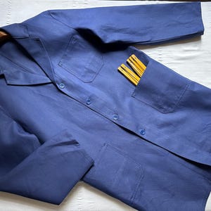 May include: A solid blue work coat featuring a collared neckline, long sleeves, and a button-down front. The coat includes one chest pocket and two lower pockets, with a yellow folding ruler partially tucked into the upper left pocket.