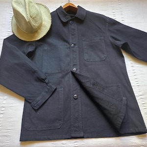 May include: A dark navy blue work jacket with a button-up front, two chest pockets, and two lower pockets. A light tan straw hat rests on the jacket. The jacket is made of a durable fabric.
