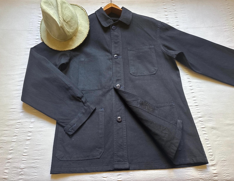 May include: A dark navy blue work jacket with a button-up front, two chest pockets, and two lower pockets. A light tan straw hat rests on the jacket. The jacket is made of a durable fabric.