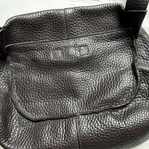 May include: A dark brown leather crossbody bag with a textured surface. The bag has a flap closure and an adjustable strap. The bag is designed with a rounded shape and a front pocket.