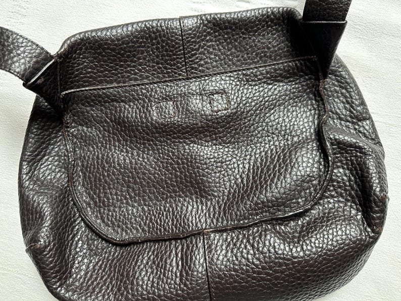 May include: A dark brown leather crossbody bag with a textured surface. The bag has a flap closure and an adjustable strap. The bag is designed with a rounded shape and a front pocket.