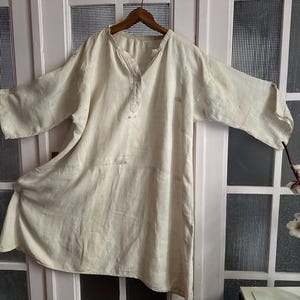 Puede incluir: Túnica de lino blanquecino con cuello en V y mangas largas y anchas. La túnica cuelga de una percha de madera contra una puerta blanca con paneles de vidrio. La tela parece ligera y ligeramente texturizada.
