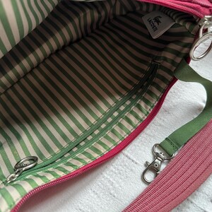 May include: Pink crossbody bag with a green and white striped interior. The bag has a green strap and a pink, ribbed shoulder strap. A small white tag with the Lacoste logo is visible.