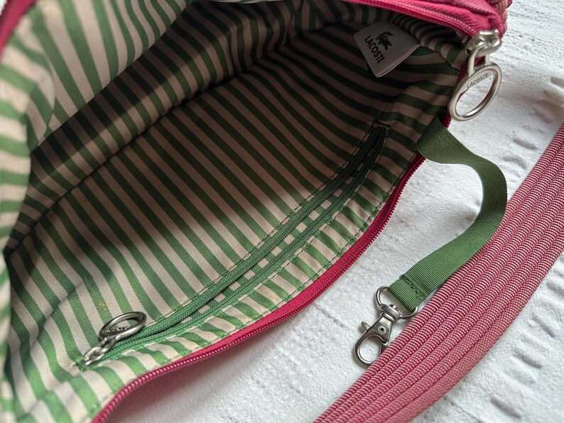 May include: Pink crossbody bag with a green and white striped interior. The bag has a green strap and a pink, ribbed shoulder strap. A small white tag with the Lacoste logo is visible.