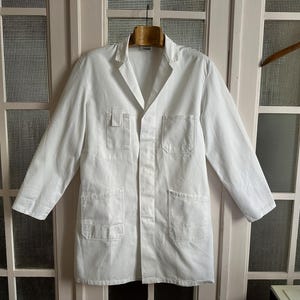 May include: A solid white lab coat featuring a notched lapel collar, long sleeves, and a button-front closure. The garment includes one chest pocket with a small vertical tab detail and two large patch pockets at the lower front section.
