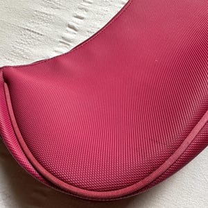 May include: A deep magenta-colored handbag with a curved, saddlebag-like shape. The bag has a textured, ribbed fabric and a matching strap. The bag's design is simple and elegant, with a focus on the rich color and material.