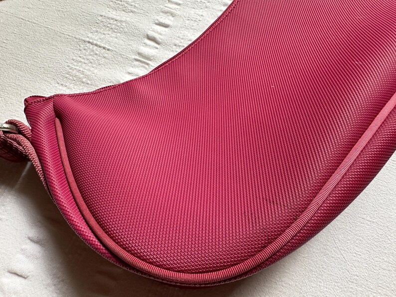 May include: A deep magenta-colored handbag with a curved, saddlebag-like shape. The bag has a textured, ribbed fabric and a matching strap. The bag's design is simple and elegant, with a focus on the rich color and material.