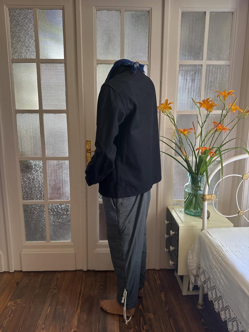 May include: A black jacket and gray pants are worn by a person. The jacket has a collar and the pants have a drawstring at the ankle. The person is standing in front of a white door with glass panes. The floor is made of wood.