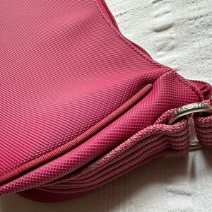 May include: Close-up of a pink handbag with a textured fabric. The bag features a rounded shape, a zipper, and a strap. A silver-colored metal ring with the brand name is visible.