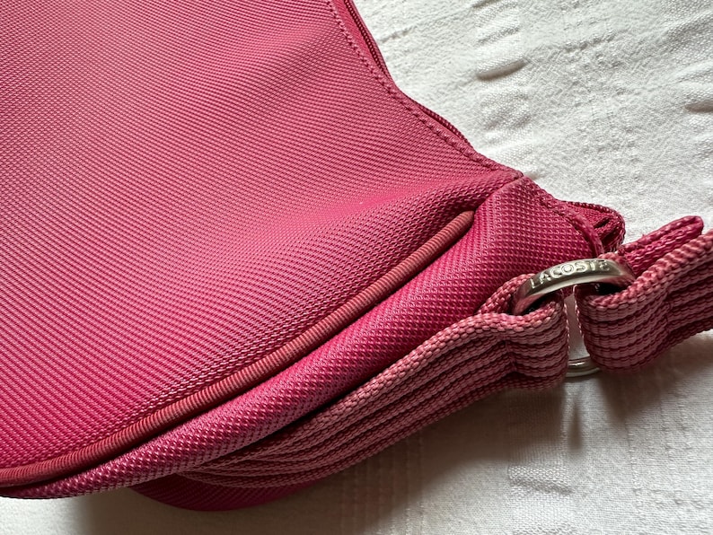 May include: Close-up of a pink handbag with a textured fabric. The bag features a rounded shape, a zipper, and a strap. A silver-colored metal ring with the brand name is visible.