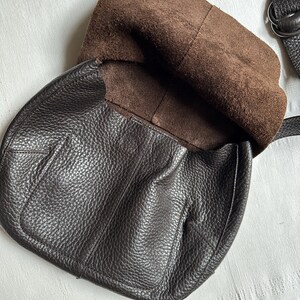 May include: Dark brown leather crossbody bag with a textured exterior. The bag has a flap closure and a brown suede interior. A dark brown strap is attached to the bag.