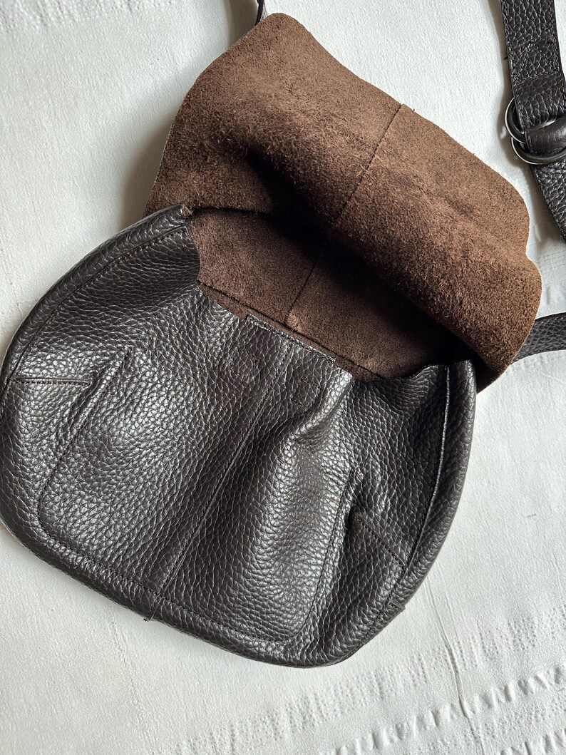 May include: Dark brown leather crossbody bag with a textured exterior. The bag has a flap closure and a brown suede interior. A dark brown strap is attached to the bag.