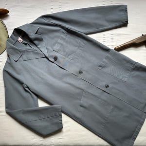 May include: A solid grey long-sleeve coat featuring a collared neckline, a button-down front closure, one chest pocket, and two lower front pockets. The coat is made of a smooth fabric and is displayed alongside a tan hat and a wooden tool.